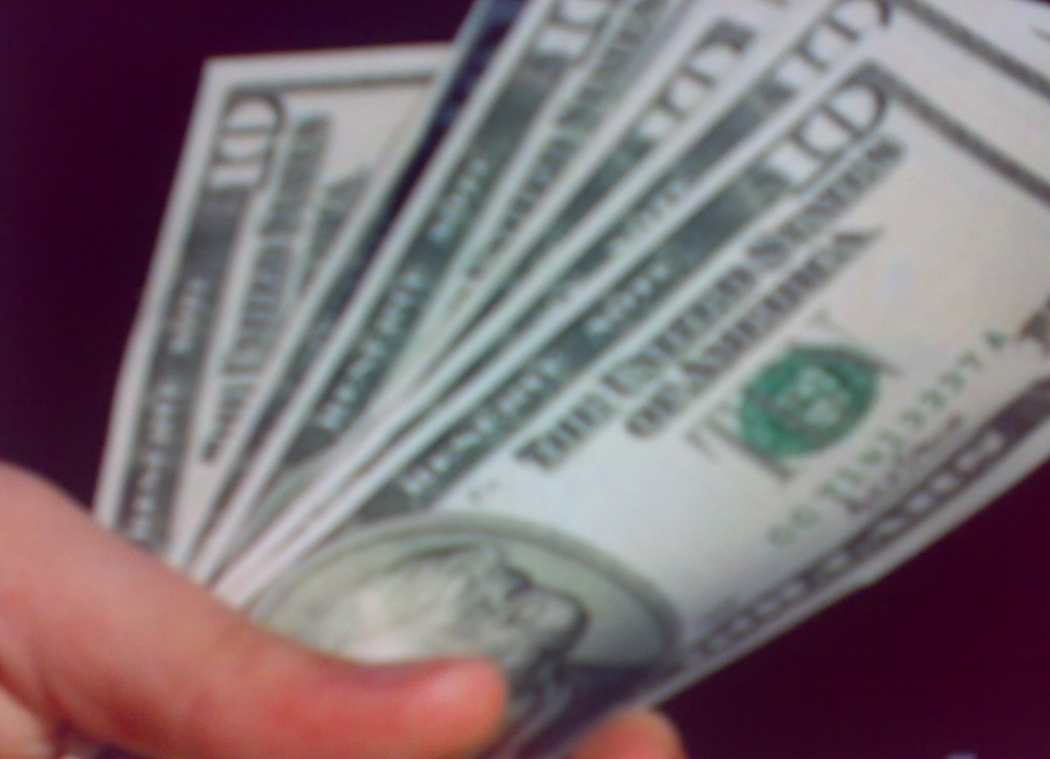 A hand holding a fanned-out stack of U.S. hundred dollar bills against a dark background.
