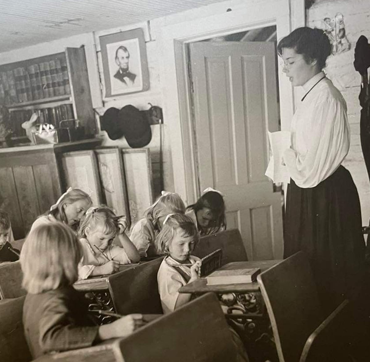 A black-and-white photo of a woman teaching young children in a classroom. The children sit at wooden desks writing on slates, while the teacher stands holding papers. Shelves, books, and a portrait hang on the wall behind them.