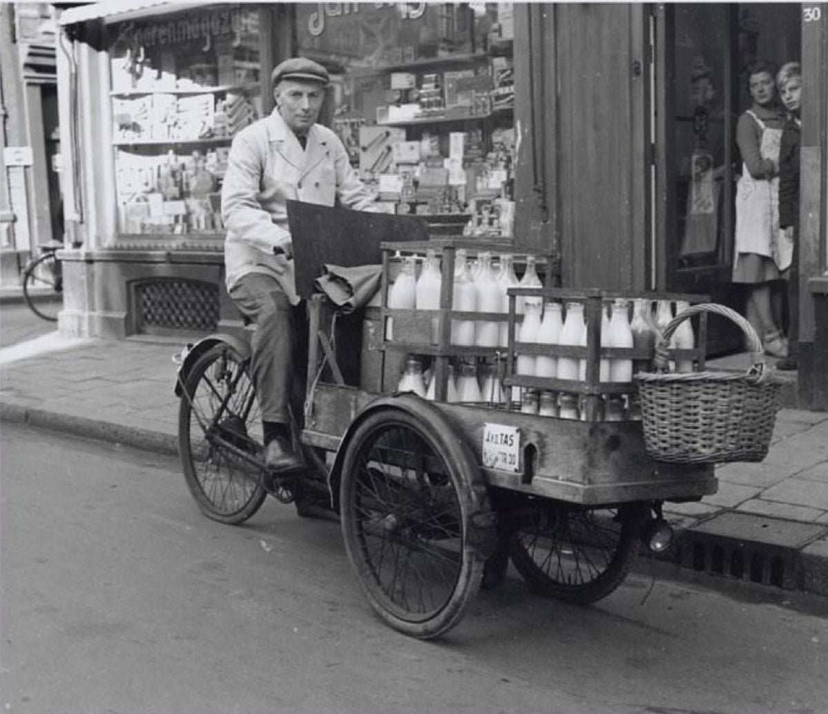 A black-and-white photo of a man riding a three-wheeled bicycle with crates of milk bottles and a basket in front, on a street with shops; two women stand in a doorway watching.