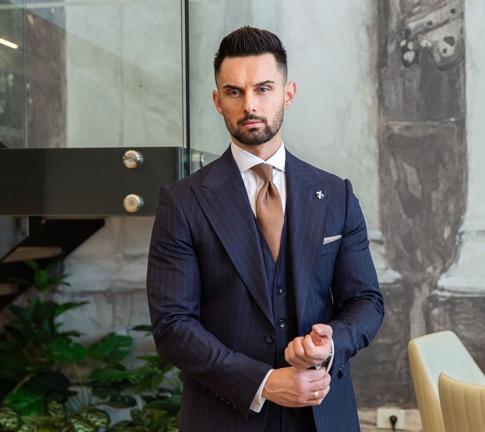 A man in a navy pinstripe suit with a tan tie stands indoors, adjusting his cuff. He has short dark hair and a beard, and is positioned near glass stairs, plants, and a wall with abstract art.