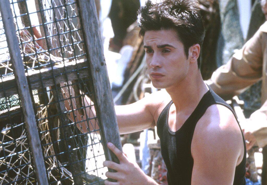 A young man with short dark hair wearing a sleeveless black shirt stands outdoors, holding onto a wooden and metal cage, looking pensively to the side.
