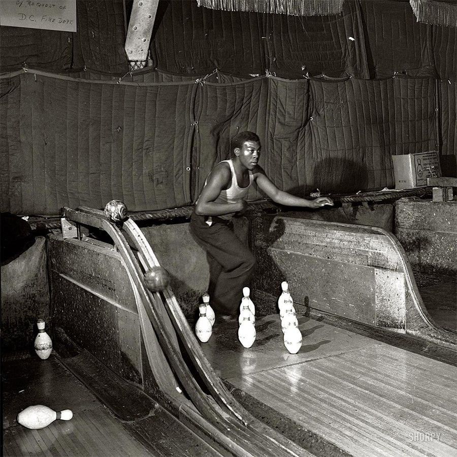 A young man wearing a tank top and pants bowls indoors, watching the ball roll down a narrow lane toward a set of ten pins. The dimly lit room has padded walls and a rustic, informal atmosphere.