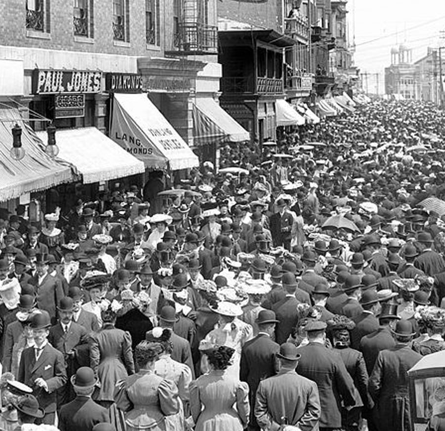 A large crowd of people in early 1900s attire, including suits, hats, and dresses, fills a city street lined with storefronts and awnings on a busy day.