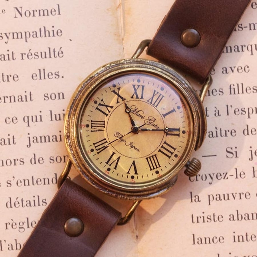 A vintage wristwatch with a brown leather strap sits on an open book. The watch face features Roman numerals and ornate clock hands, with cursive writing at the center. The book beneath has French text.