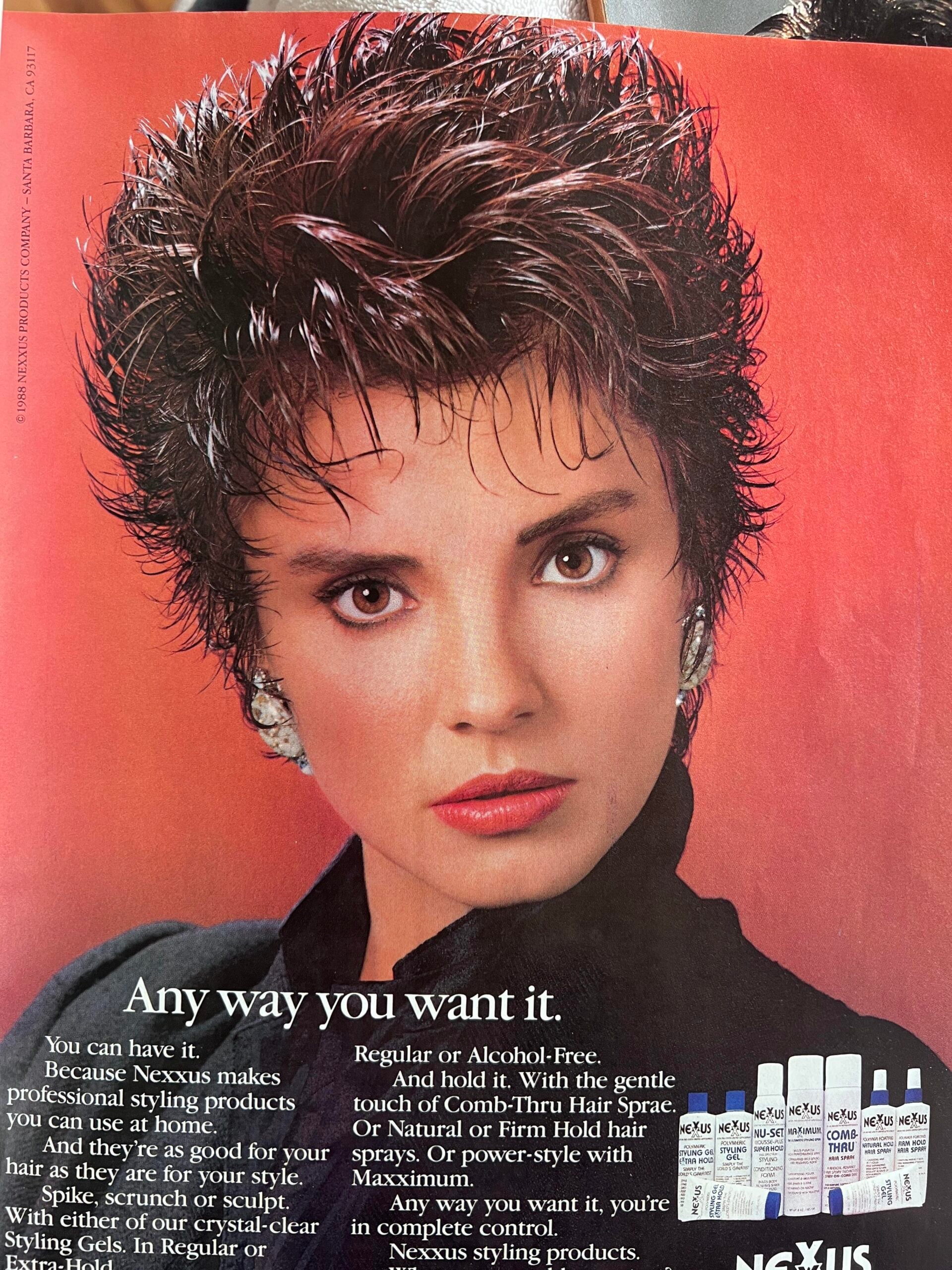 A woman with short, spiky dark hair and bold makeup poses against a red background. She wears silver earrings and a black top. At the bottom, there are several Nexxus hair styling products displayed and text about them.