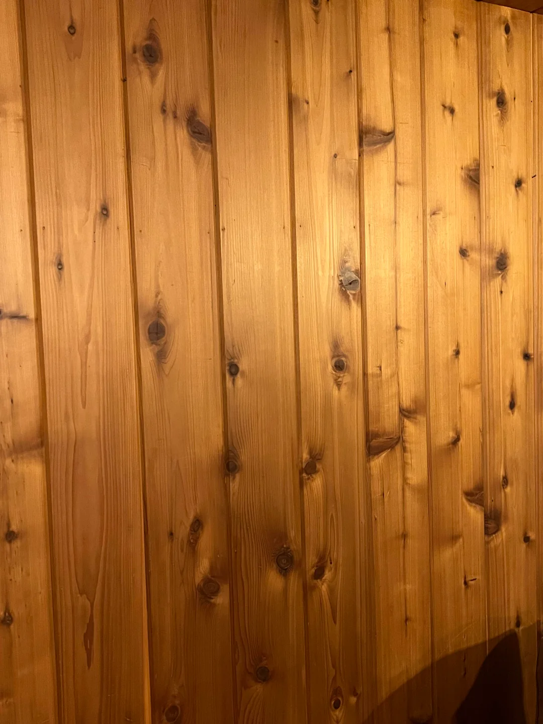 Wooden wall with vertical natural planks, featuring visible wood grain, knots, and warm brown tones. The lighting highlights the texture and variations in the wood surface.
