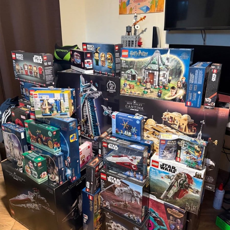 A large collection of LEGO sets, mainly Star Wars and Harry Potter themes, stacked on the floor in front of a TV and a brown curtain. Some sets are large and unopened, with colorful boxes on display.