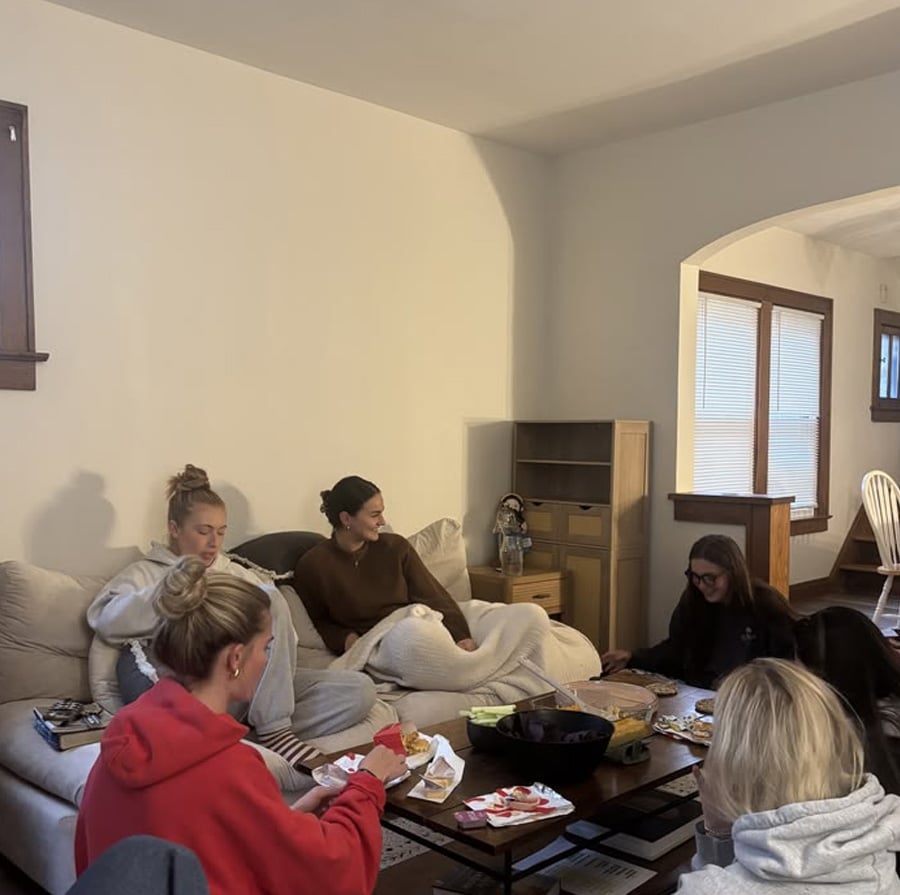 Six women sit together in a cozy living room, eating snacks and chatting. Some are seated on a couch with blankets, while others are on the floor around a coffee table filled with food. The atmosphere is warm and relaxed.