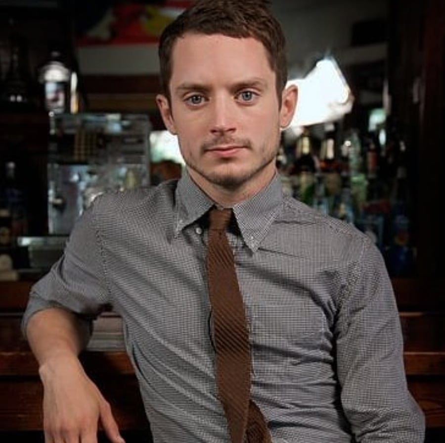 A man with short brown hair and a trimmed beard sits at a bar, wearing a grey checkered shirt and a brown tie, looking directly at the camera with a neutral expression.