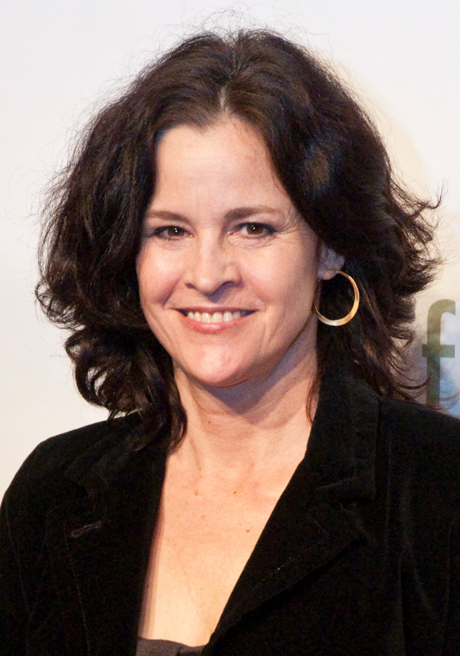 A woman with wavy dark hair wearing gold hoop earrings and a black blazer smiles at the camera against a light background.