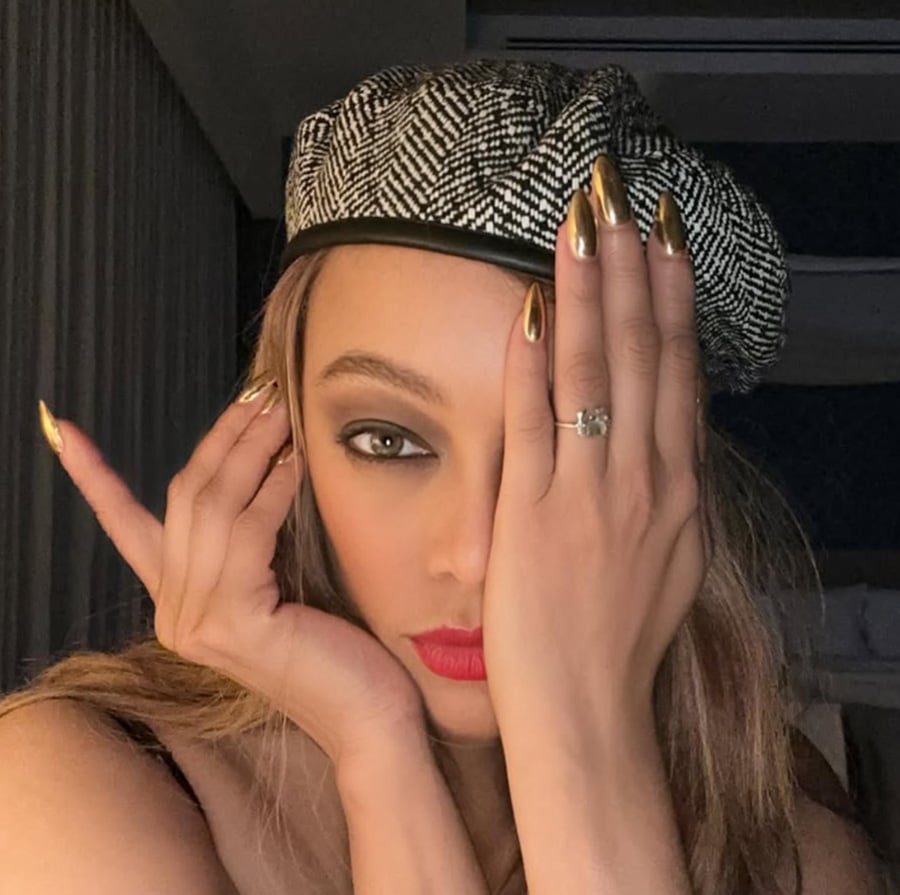 A woman with long, gold-painted nails covers one eye with her hand, wearing red lipstick and a patterned beret, and looking directly at the camera. She has a ring on her finger and dramatic eye makeup.