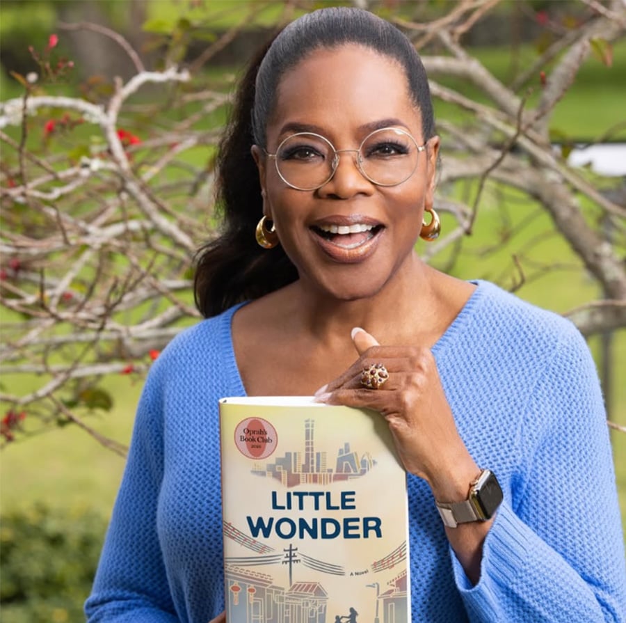 A woman with glasses and a blue sweater smiles outdoors while holding a book titled "Little Wonder." She has dark hair pulled back and wears hoop earrings, a ring, and a watch. There's greenery and branches in the background.