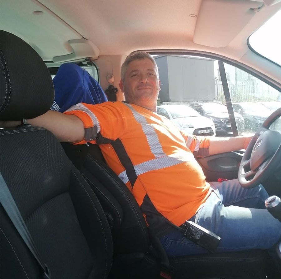 A man wearing a bright orange high-visibility shirt sits in the driver's seat of a vehicle, resting his arm on the backrest and smiling at the camera. Sunlight shines through the window.