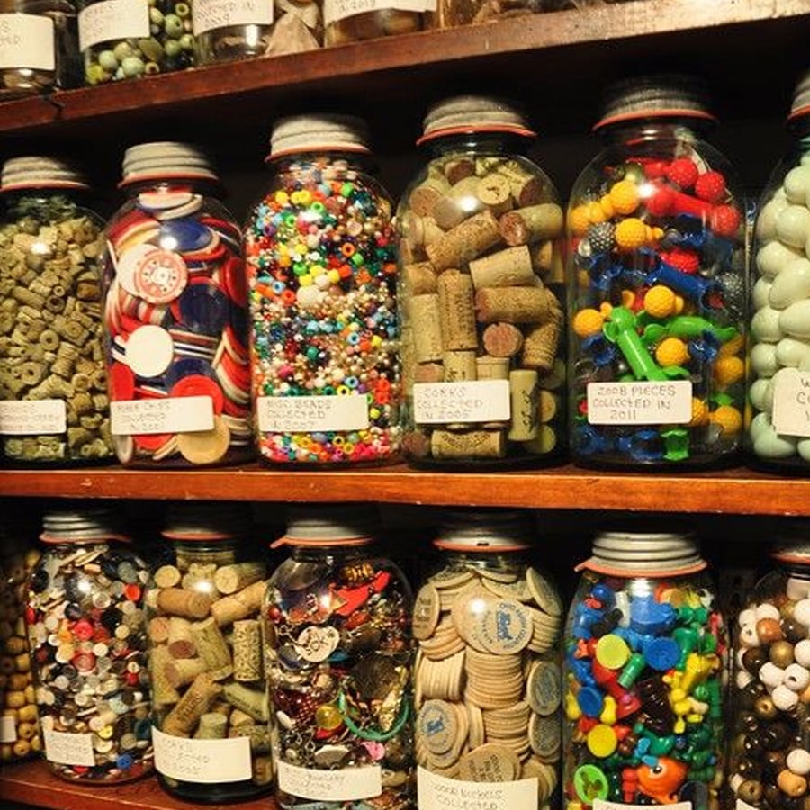 Glass jars filled with various colorful objects like beads, buttons, corks, and other craft materials are neatly arranged on wooden shelves, each jar labeled with handwritten tags.