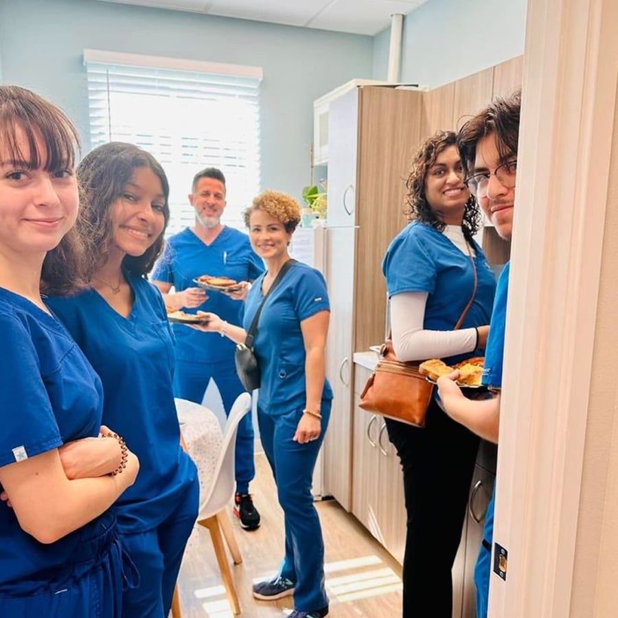 A group of six people in blue medical scrubs smile and stand together in a break room, holding plates of food. They appear relaxed and happy, with natural light coming in through a window in the background.