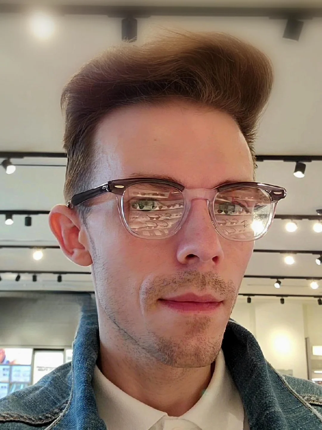 A man with light skin, short brown hair styled upwards, and glasses with clear frames is wearing a denim jacket and white shirt, standing indoors under ceiling lights, with reflections visible in his glasses.