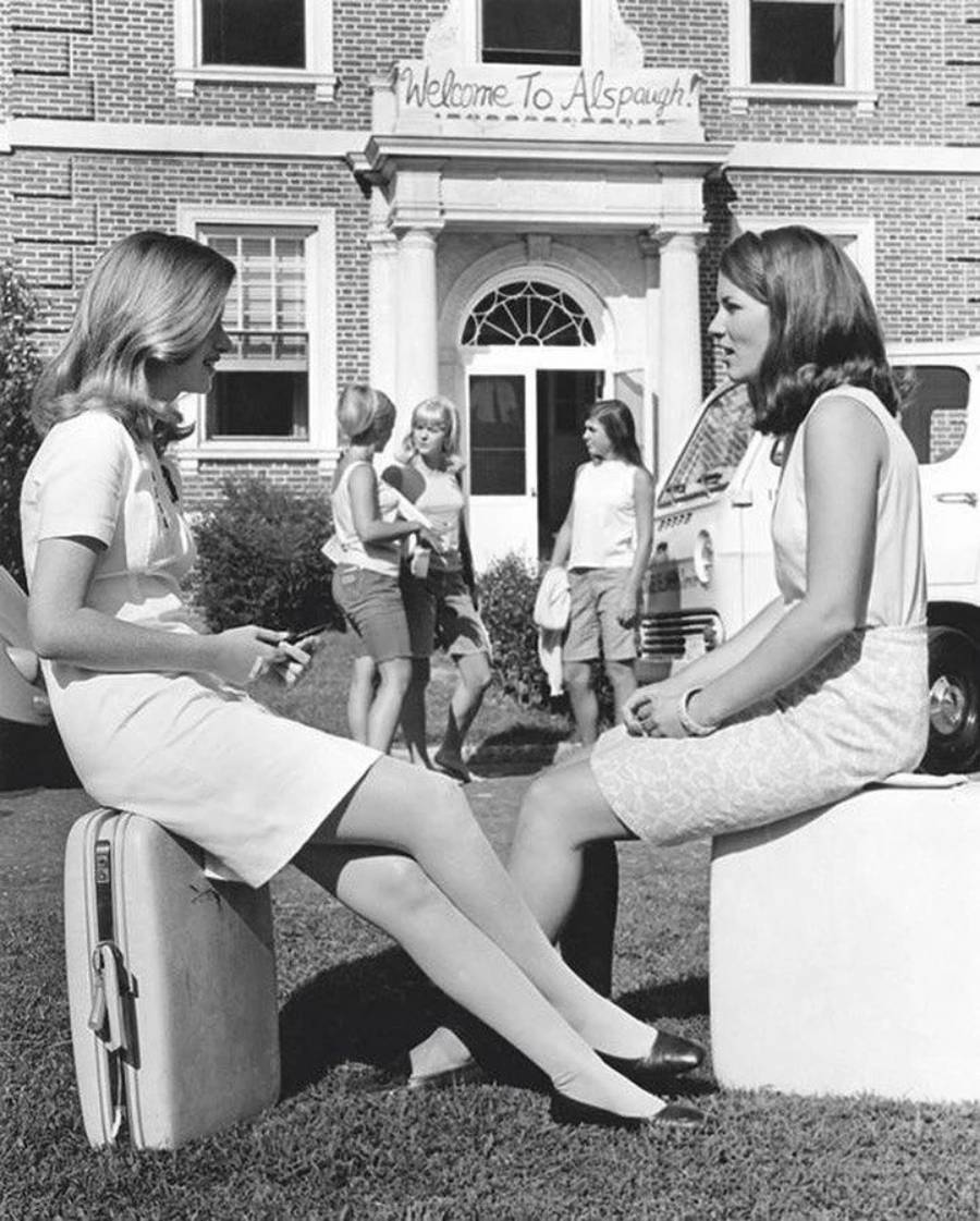 Two young women sit on suitcases in front of a building with a "Welcome to Alspaugh" sign, while three other women are chatting and walking in the background. A van is parked nearby. The scene appears to be from the 1960s.