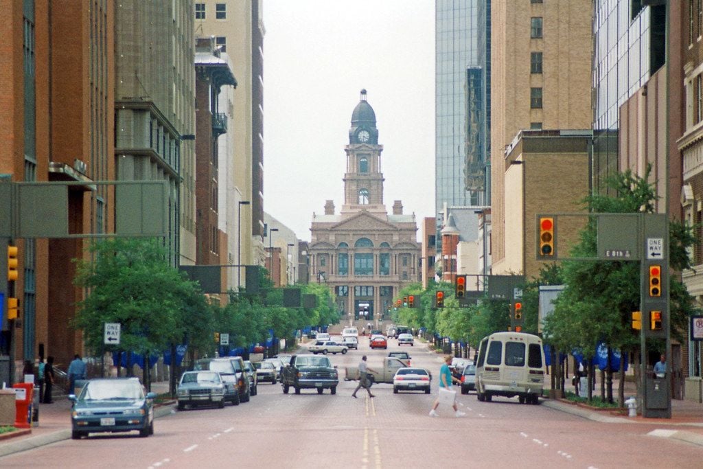 A busy city street with cars, pedestrians crossing, and tall buildings on both sides, leading to a historic courthouse with a clock tower in the background. Traffic lights are visible along the road.