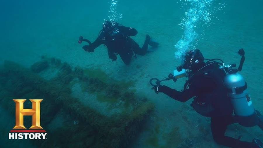 Two scuba divers explore an underwater site with overgrown ruins or artifacts. Both are in black suits with oxygen tanks. The History Channel logo appears in the bottom left corner.