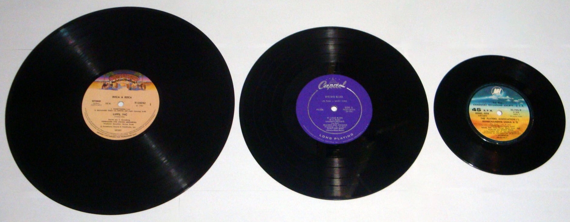 Three vinyl records of different sizes are arranged side by side on a white surface, showing their colored center labels: a 12-inch on the left, a 10-inch in the middle, and a 7-inch on the right.