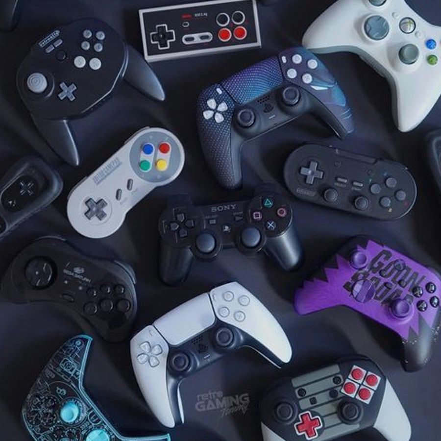 A variety of video game controllers from different consoles and eras are arranged on a flat surface, showcasing diverse shapes, colors, and button layouts.