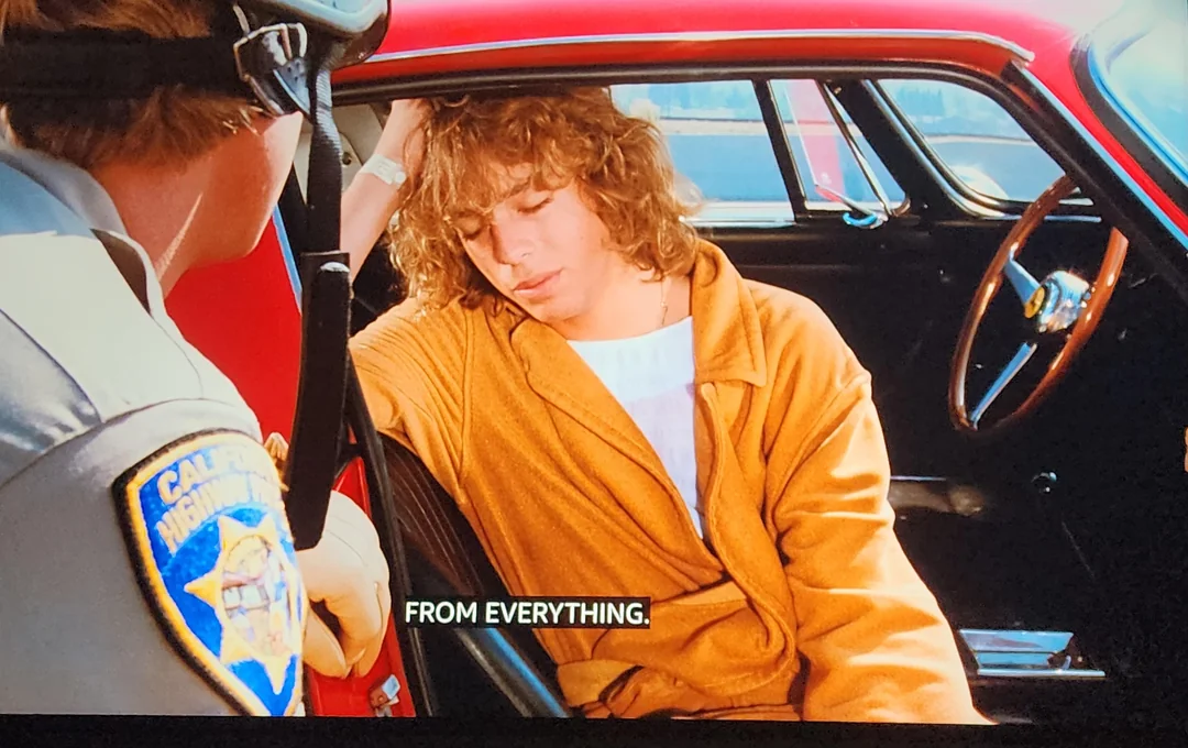 A young man with curly hair, wearing a tan robe, sits slouched in the passenger seat of a red car, eyes closed, as a uniformed California Highway Patrol officer leans in through the open door. Subtitles read: "FROM EVERYTHING.