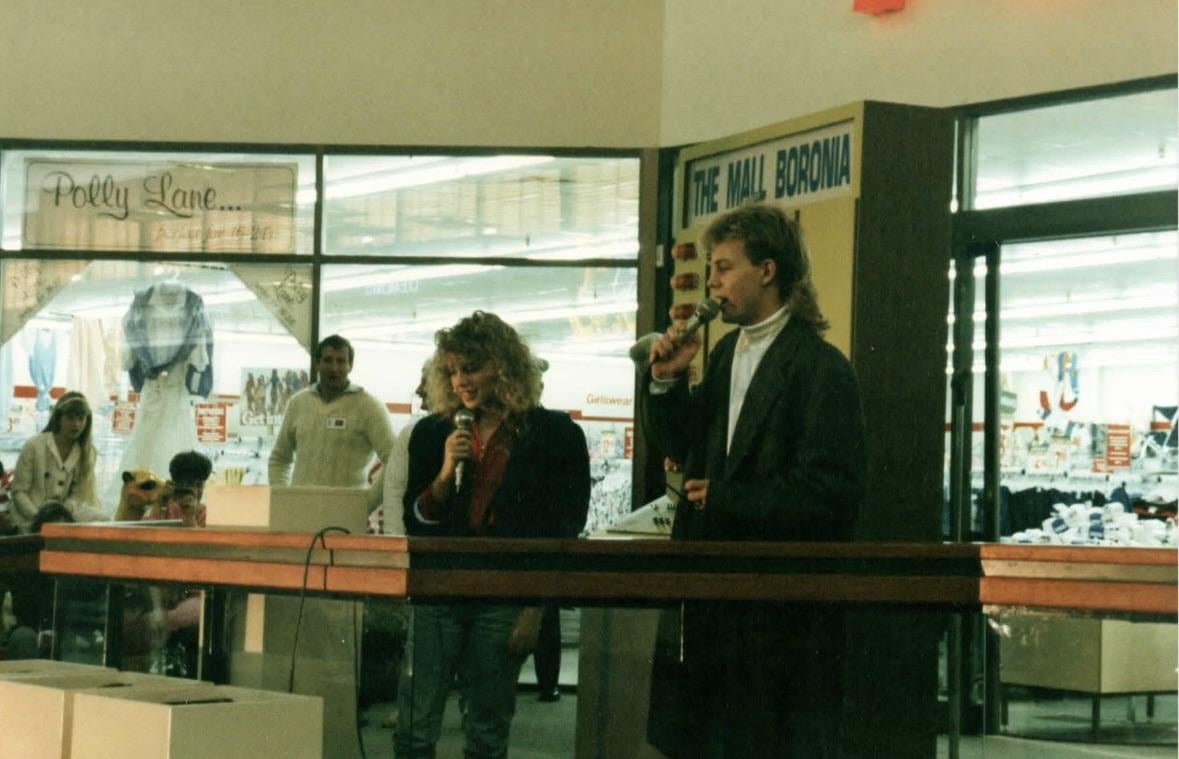Two people hold microphones and sing in a shopping mall, standing behind a red railing. Shoppers and store signs are visible in the background, with a Polly Lane store and "The Mall, Boronia" sign nearby.