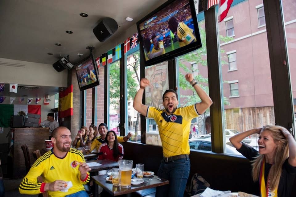 A group of people in yellow soccer jerseys celebrates and cheers inside a bar with flags and TVs showing a soccer match. One man is standing and raising his arms in excitement. Food and drinks are on the table.