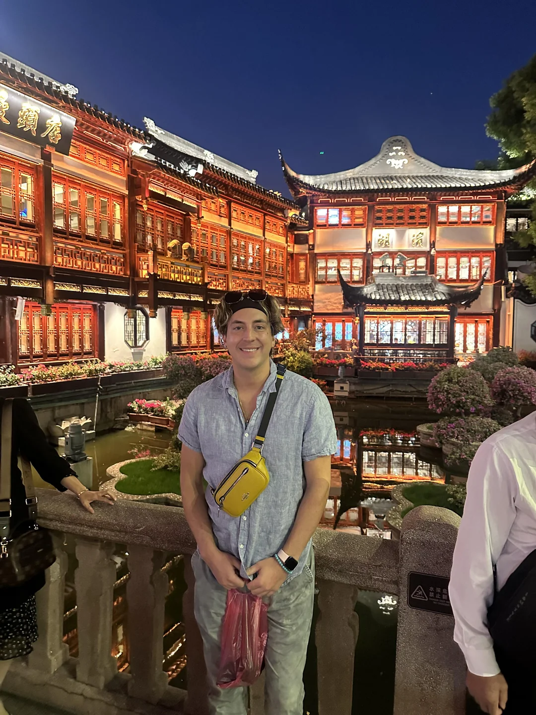 A man wearing a gray shirt, yellow fanny pack, and a black cap stands smiling on a stone bridge at night, with brightly lit traditional Chinese buildings in the background and their reflection in the water.