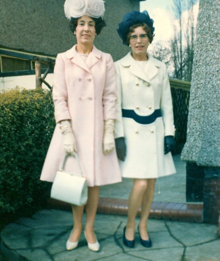 Two women stand side by side outdoors, wearing elegant 1960s-style coats and hats. One is in pink with a white purse and gloves; the other is in white with a dark belt, blue gloves, and a blue hat. Both are dressed formally.