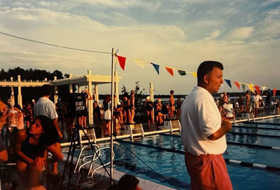 A man in a white shirt and red shorts stands by a swimming pool, holding a drink. Behind him, people gather near the pool, some seated, with colorful pennant flags strung overhead under a partly cloudy sky.