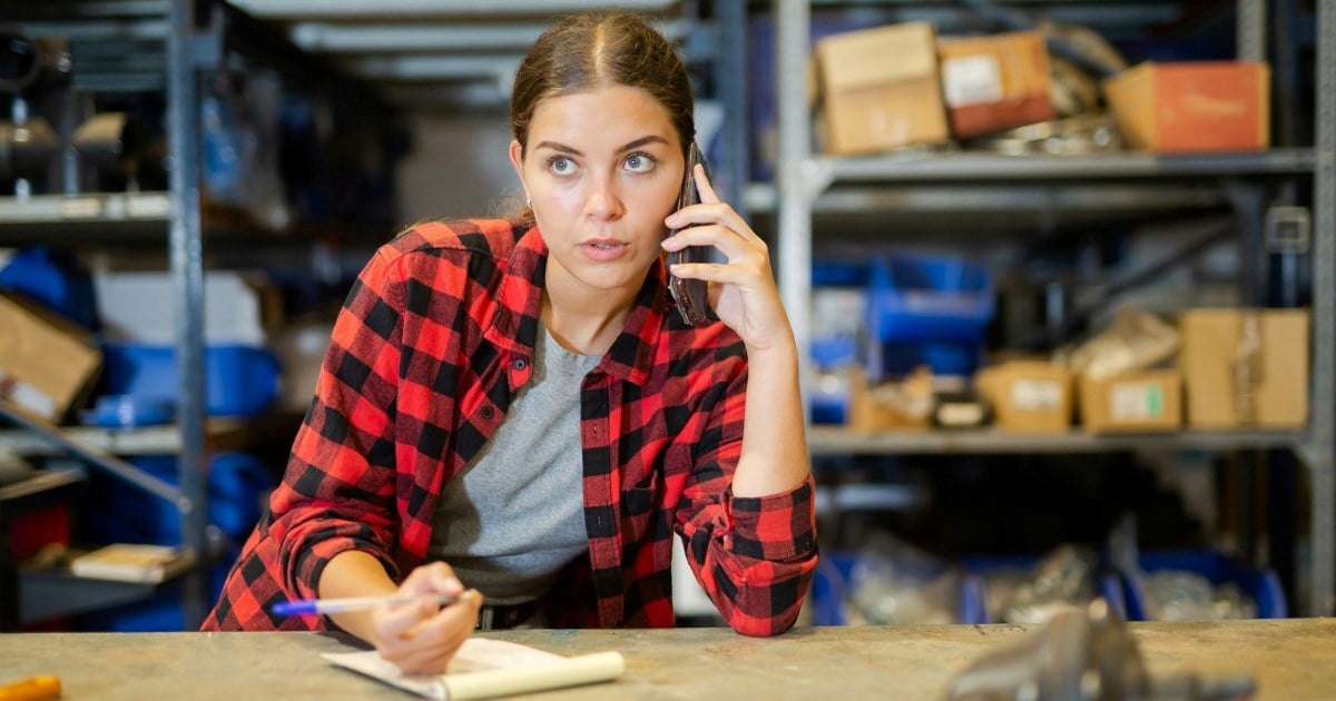 A woman in a red plaid shirt sits at a workbench in a warehouse, talking on her phone while writing in a notebook, surrounded by shelves filled with boxes and tools.