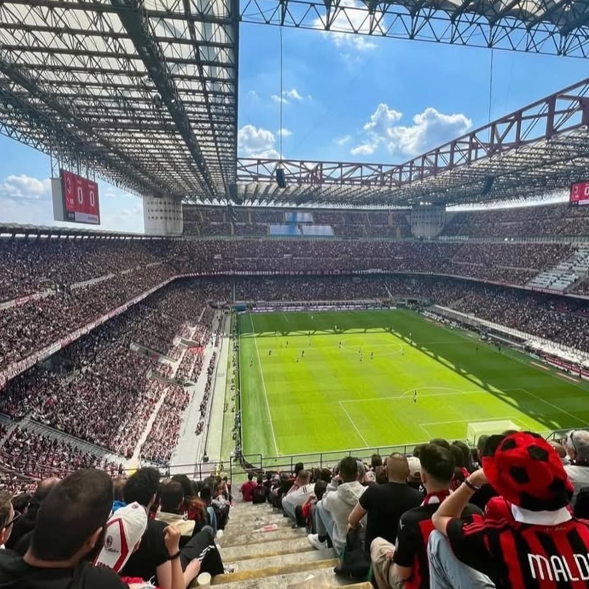 View from the stands of a packed football stadium during a daytime match, with fans in red and black jerseys watching the game on a bright green field under a partly cloudy sky.