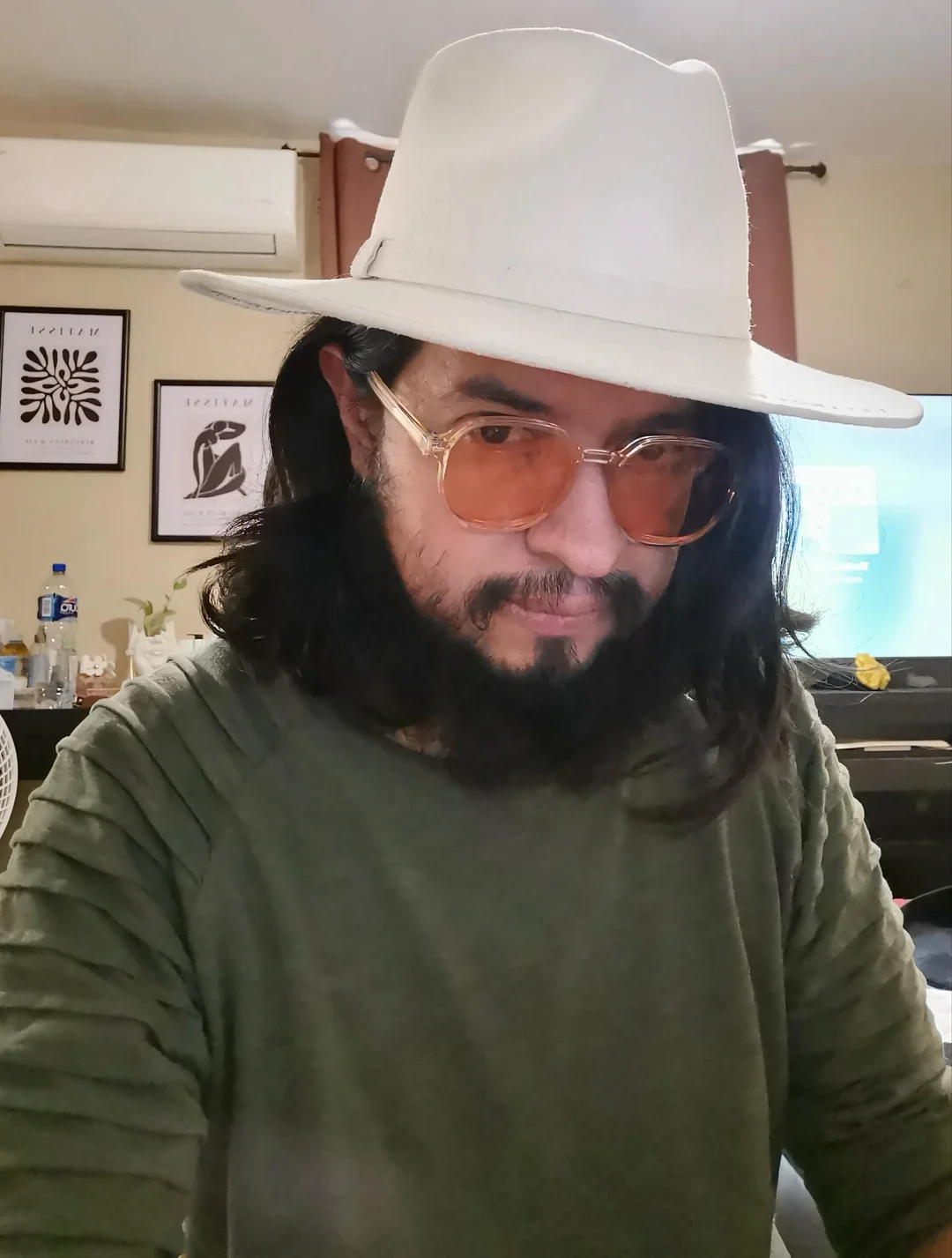 A person with long dark hair, a beard, and mustache wears a white hat, tinted glasses, and a green shirt, sitting indoors with framed art, a TV, and various items in the background.