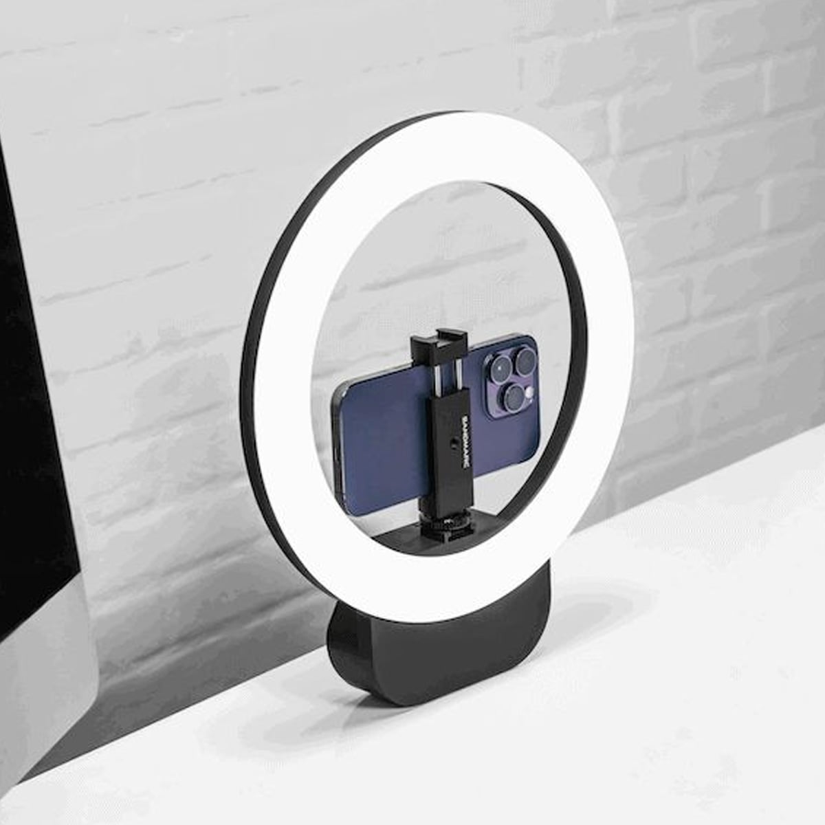 A smartphone is held vertically in a mount at the center of a lit circular ring light, placed on a white desk near a computer monitor and a light gray brick wall.