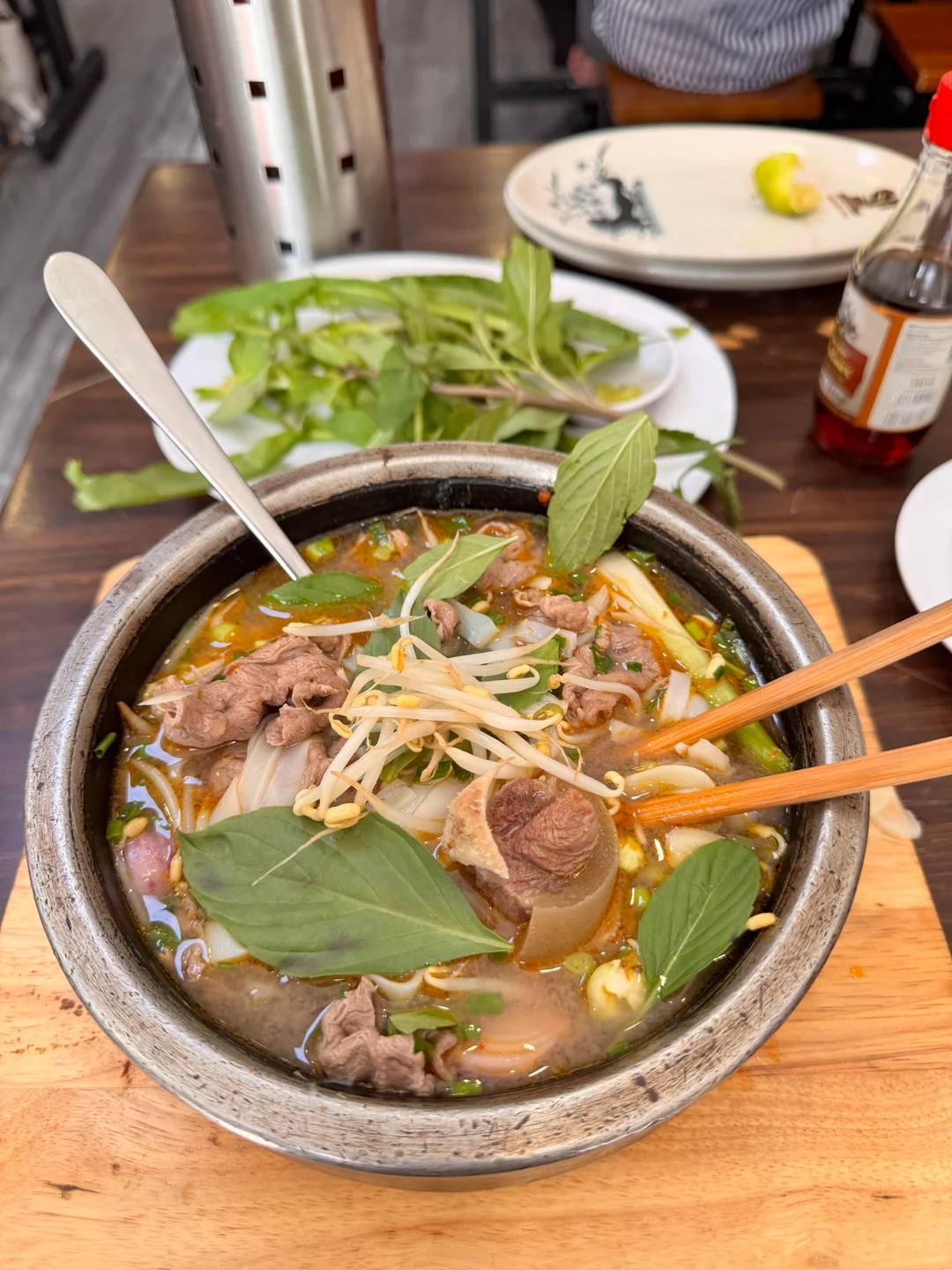 A bowl of Vietnamese noodle soup topped with beef, bone, fresh basil, and bean sprouts sits on a wooden tray. Chopsticks and a spoon are in the bowl. Fresh herbs, condiments, and plates are on the table nearby.