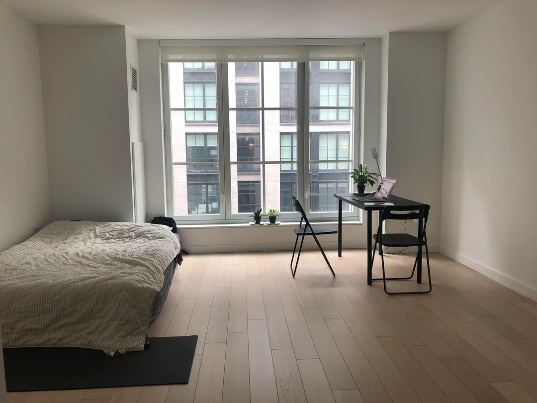 Minimalist bedroom with a bed on the left, a desk with a laptop and lamp on the right, two chairs, light wooden floor, and large windows letting in natural light with city buildings visible outside.