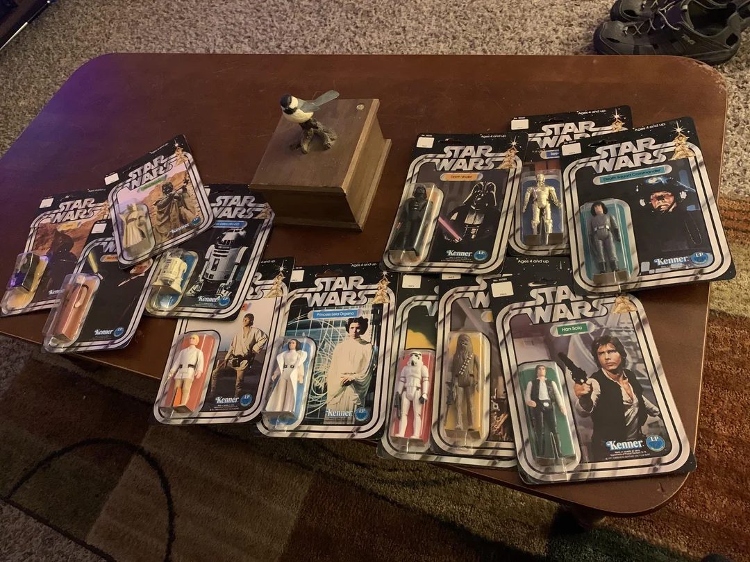 Ten vintage Star Wars action figures in original Kenner packaging are spread out on a wooden coffee table, with a small dog figurine and other items also visible on the table.