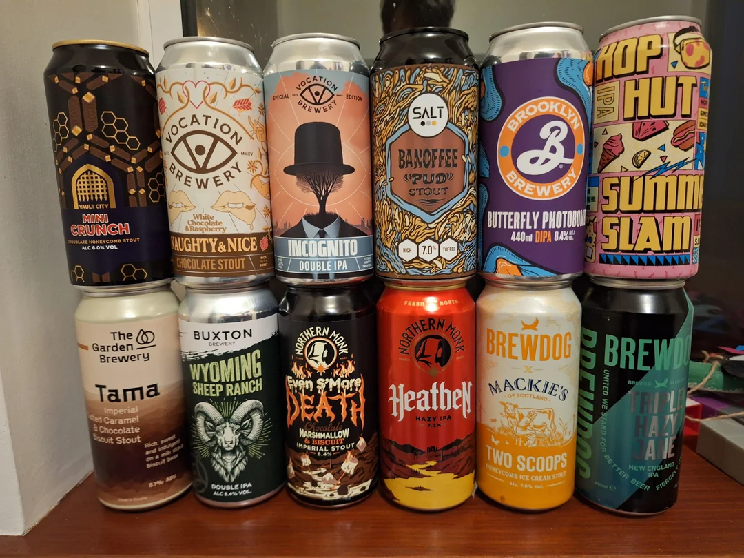 Twelve colorful craft beer cans with distinctive labels and designs are arranged in three rows on a shelf, showcasing various breweries and beer styles.