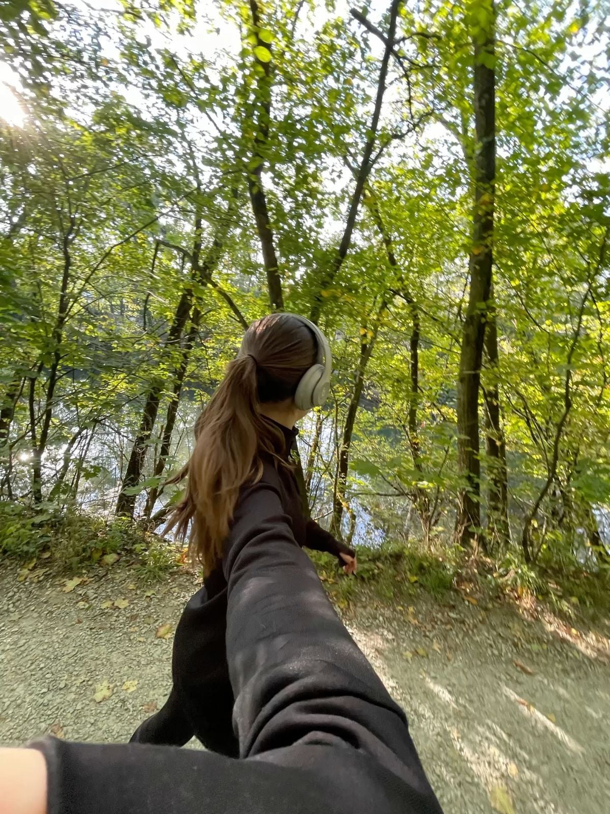 A person with long brown hair, wearing headphones and a black outfit, walks on a forest path while holding someone’s hand. Sunlight filters through green trees, and a calm river is visible in the background.