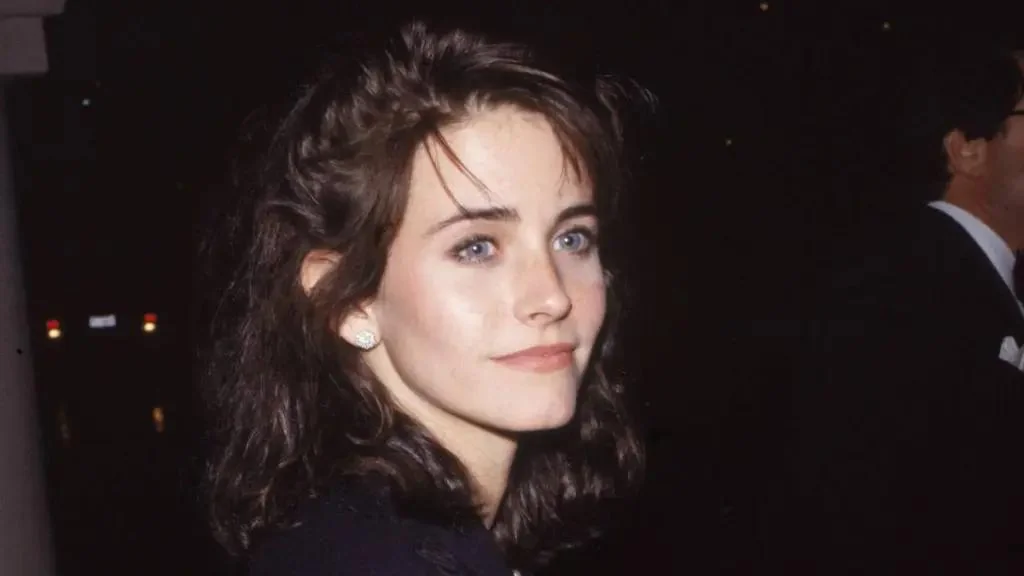 A woman with wavy dark hair and blue eyes is wearing pearl earrings and a dark top, looking slightly to the side at night. The background is dark and blurry, with another person in a suit partially visible.