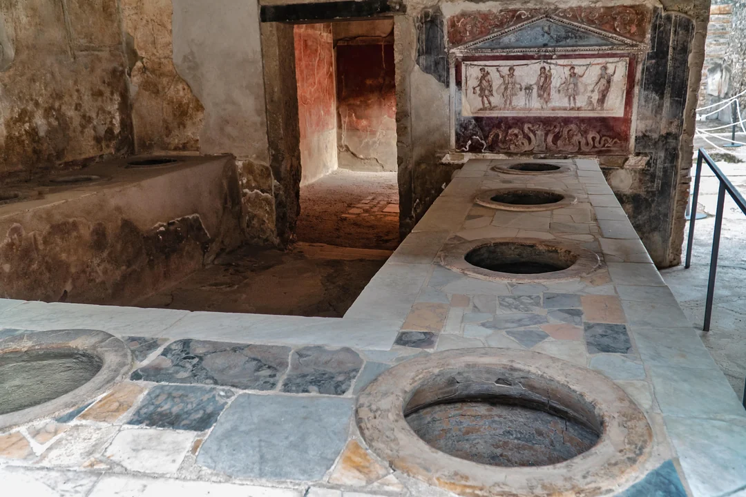 Ancient stone counters with round holes for pots in a Roman thermopolium, with faded wall paintings in the background and a doorway leading to another room.