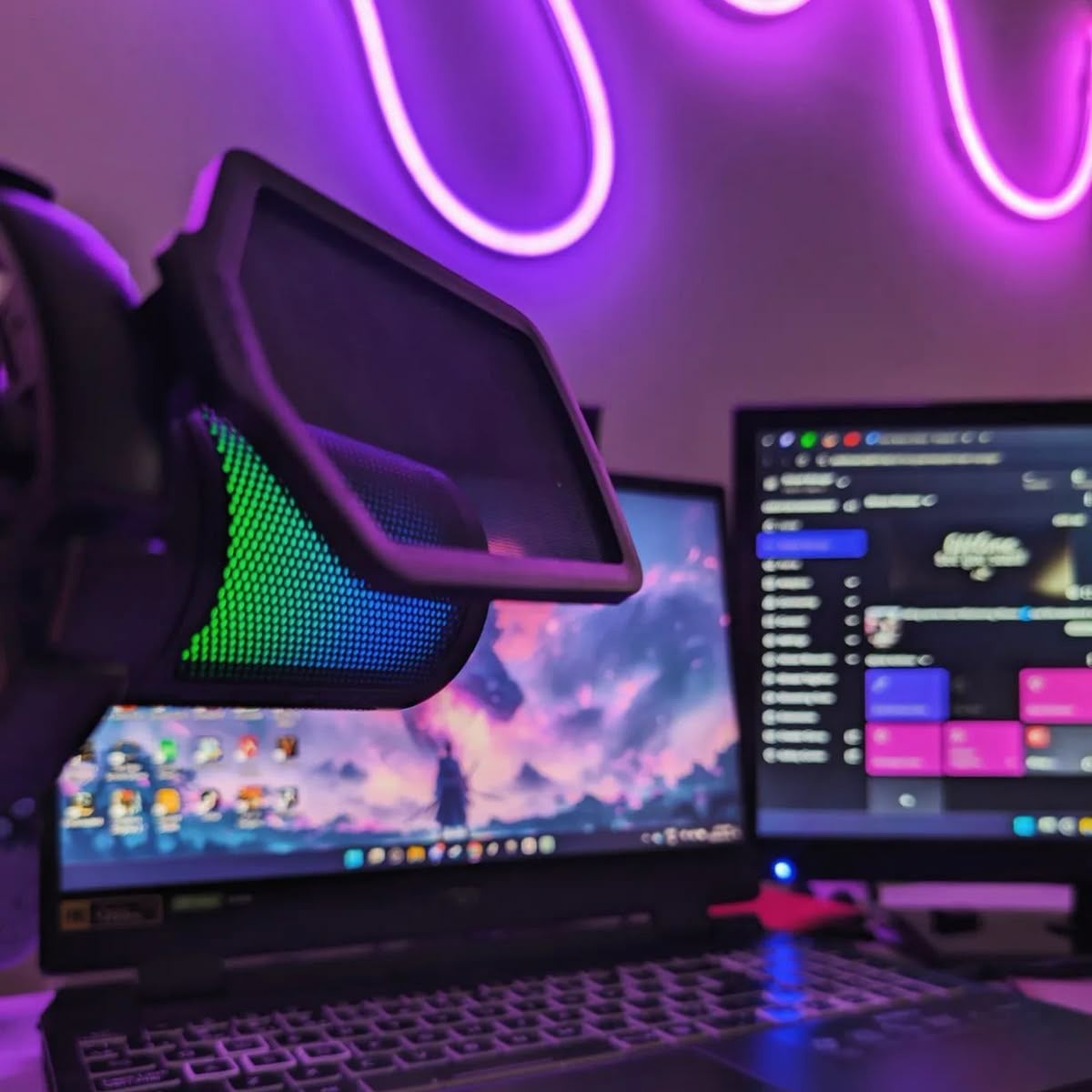 A desktop setup with a colorful RGB microphone in the foreground, a laptop displaying a fantasy landscape wallpaper, and a monitor showing a streaming dashboard. Neon purple LED lights decorate the background wall.