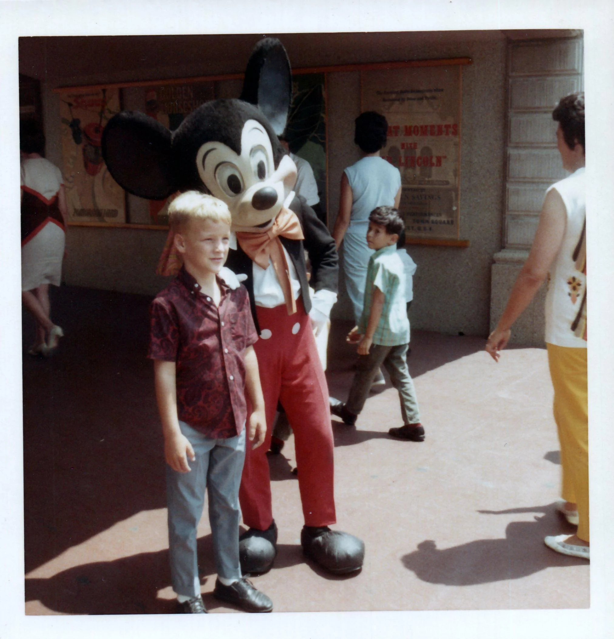 A young boy stands smiling next to a person in a classic Mickey Mouse costume at an amusement park. Several other people walk by in the background on a sunny day.