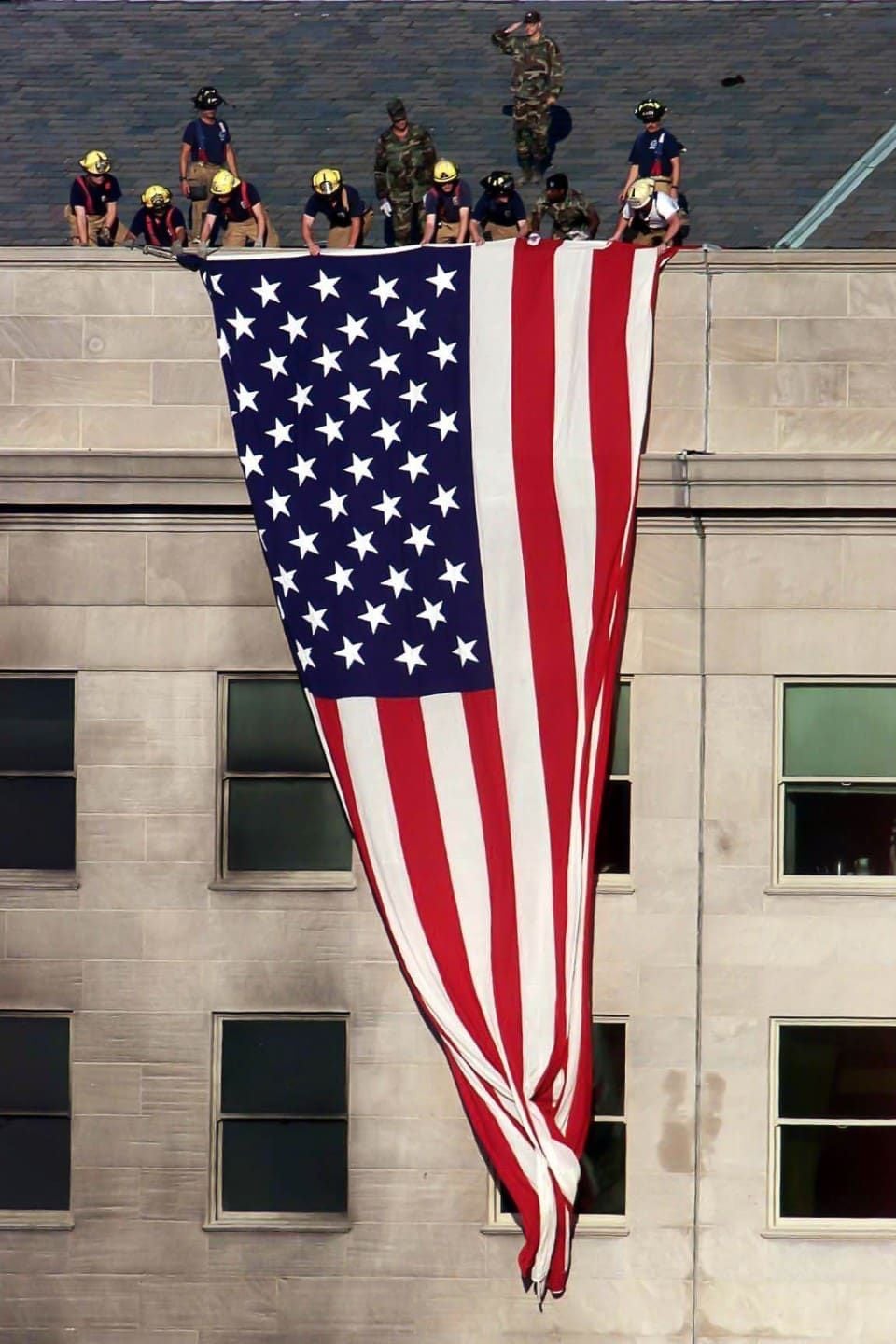 Firefighters and military personnel display a large, vertically hanging American flag over the edge of a beige building, with several windows visible below the flag.