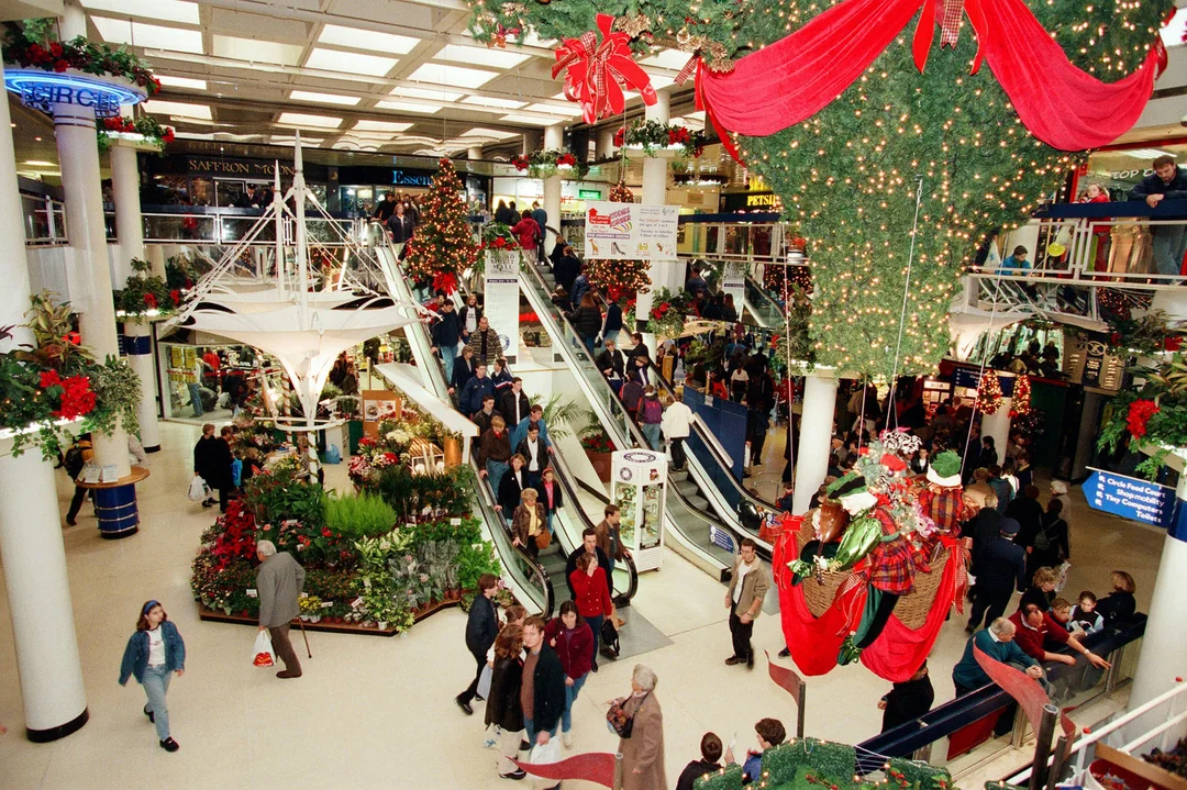 A busy shopping mall decorated with Christmas lights, garlands, ornaments, and a large upside-down Christmas tree. Shoppers walk around, some using escalators, and festive decorations hang from the ceiling.