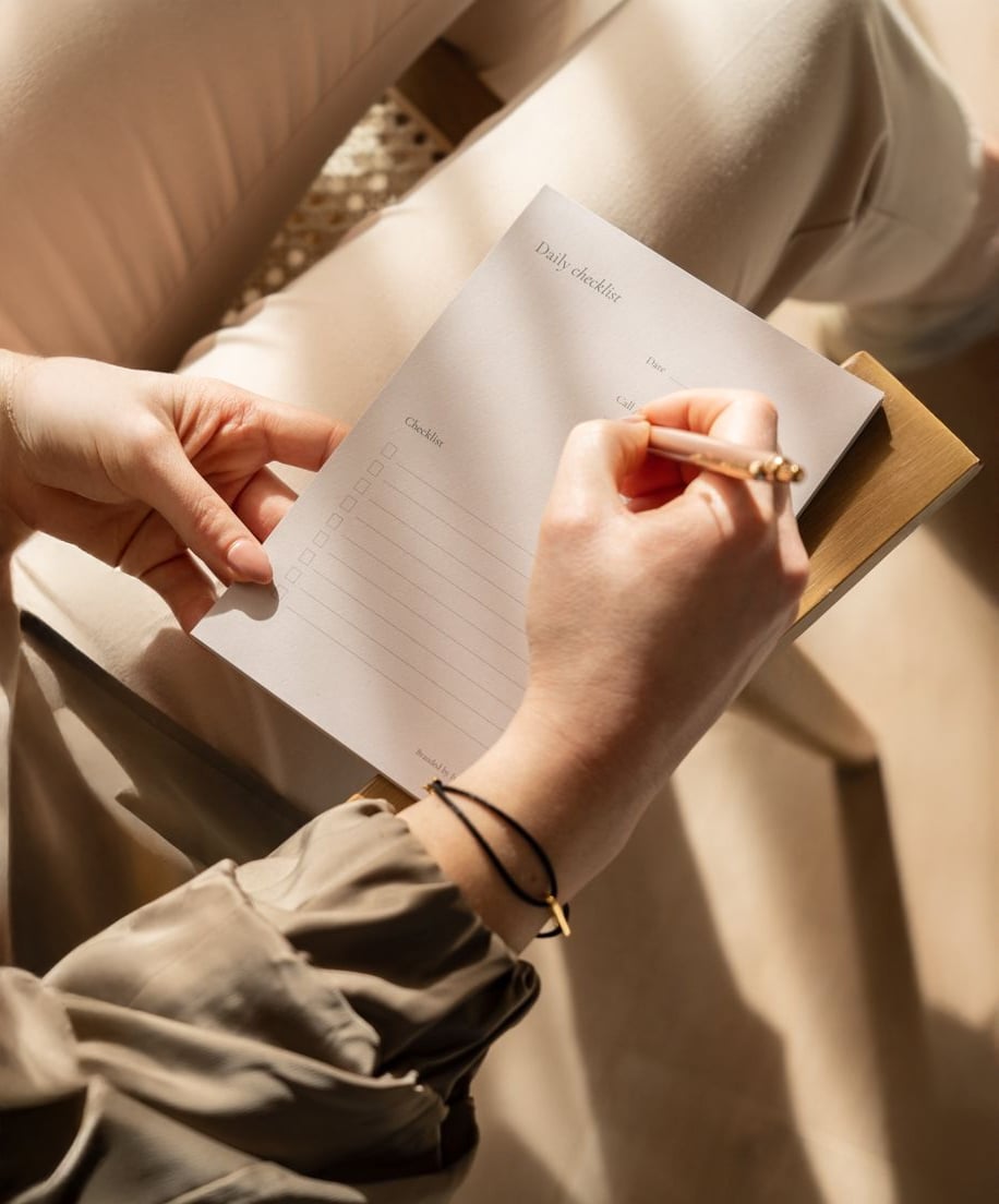 A person sits and holds a clipboard with a daily checklist, writing on it with a pencil. Sunlight filters over their hands and lap, casting soft shadows on the paper and their light-colored clothes.