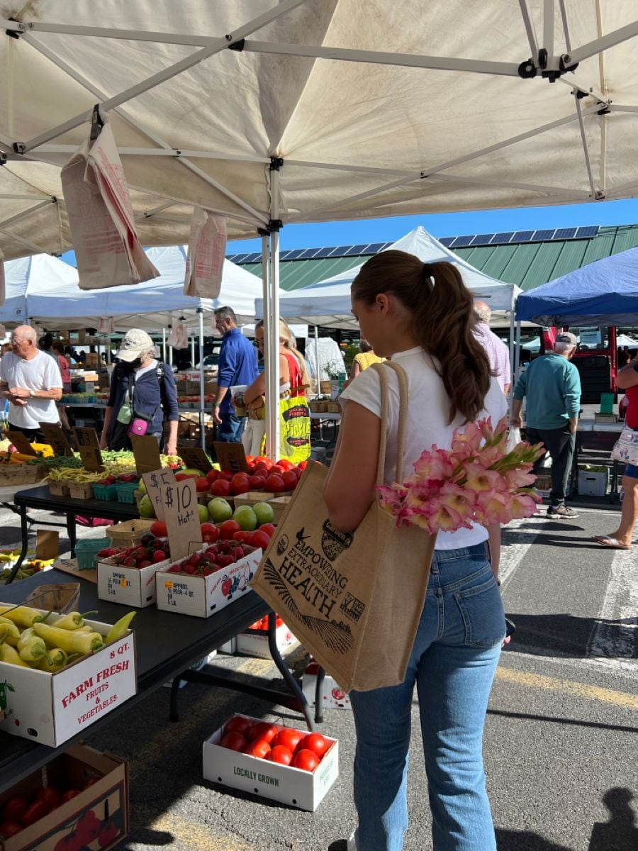 A woman with a ponytail carries a tote bag with pink flowers while shopping at an outdoor farmers market, surrounded by fruit and vegetable stands under white tents on a sunny day.