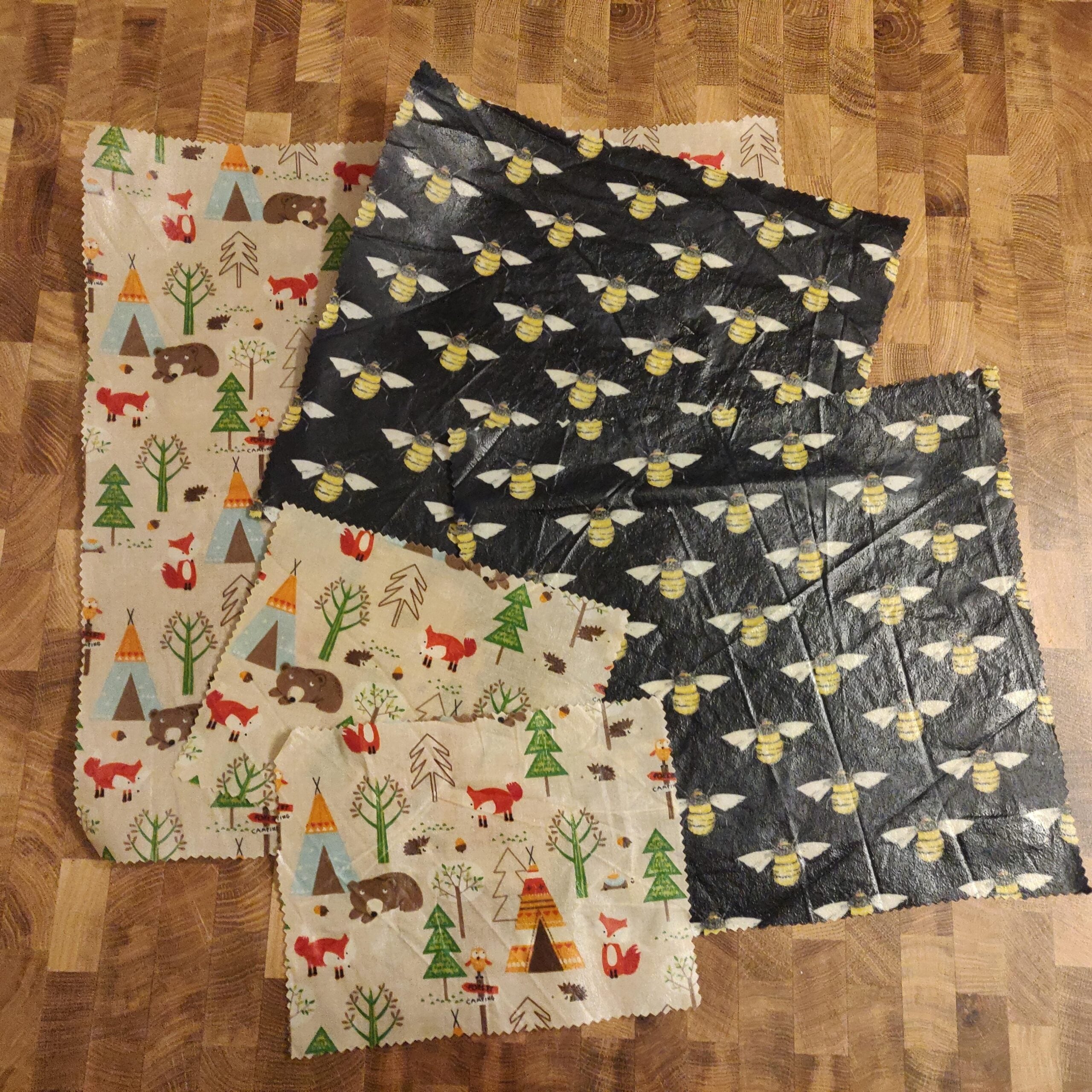 Four reusable beeswax wraps are laid out on a wooden surface. Two have a woodland animal and tree pattern, and two are black with a repeating yellow bee design.