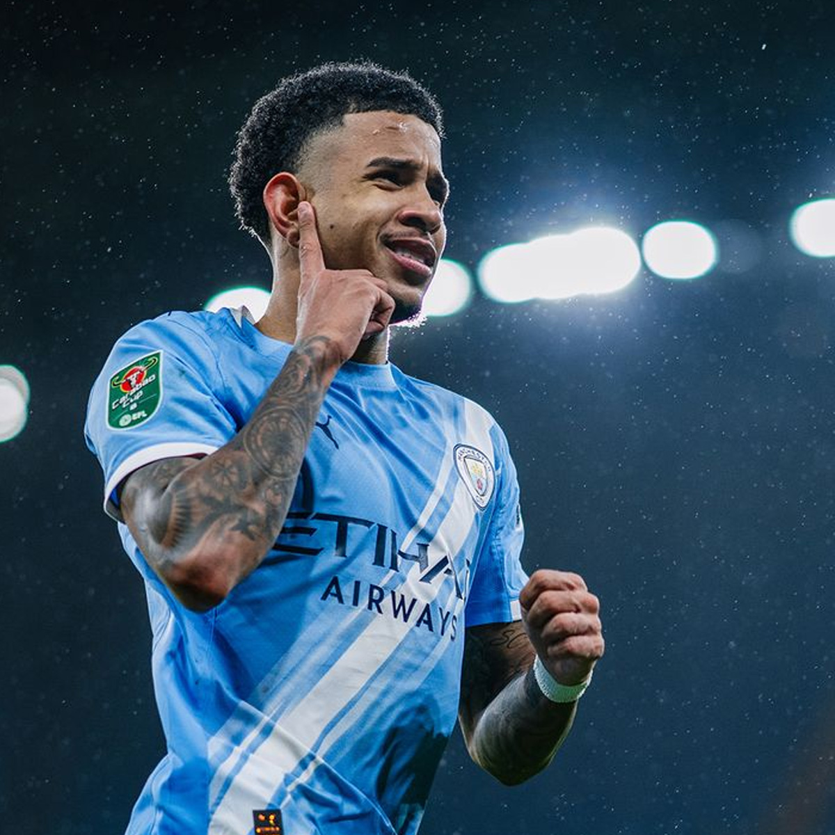 A soccer player wearing a light blue Manchester City jersey celebrates on the field, pointing to his ear with one hand and clenching his fist with the other, with bright stadium lights in the background.